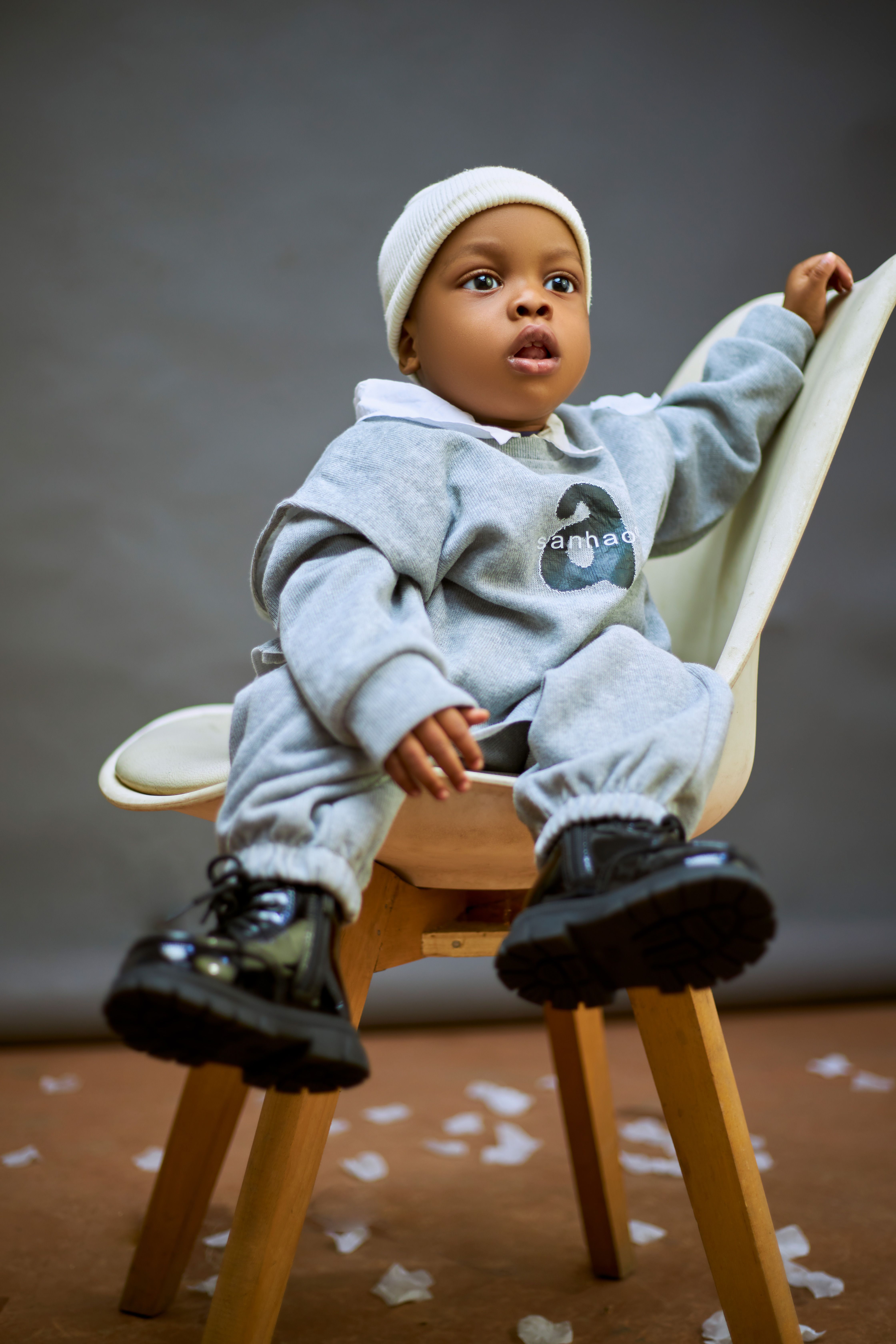 Cute baby in a casual gray outfit sitting on a modern chair with a neutral background.