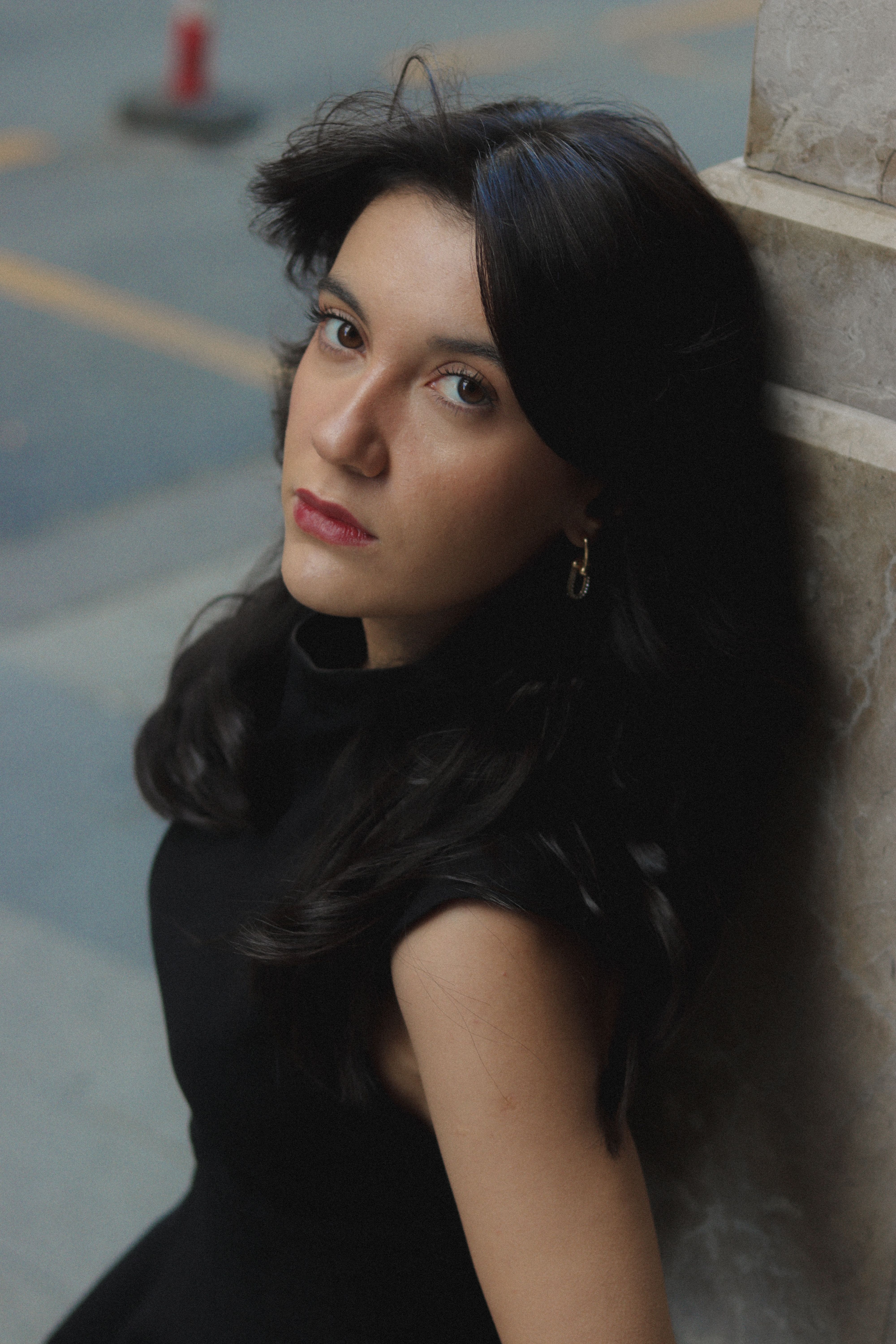 Young woman with long dark hair in black dress, leaning on marble wall with thoughtful expression.