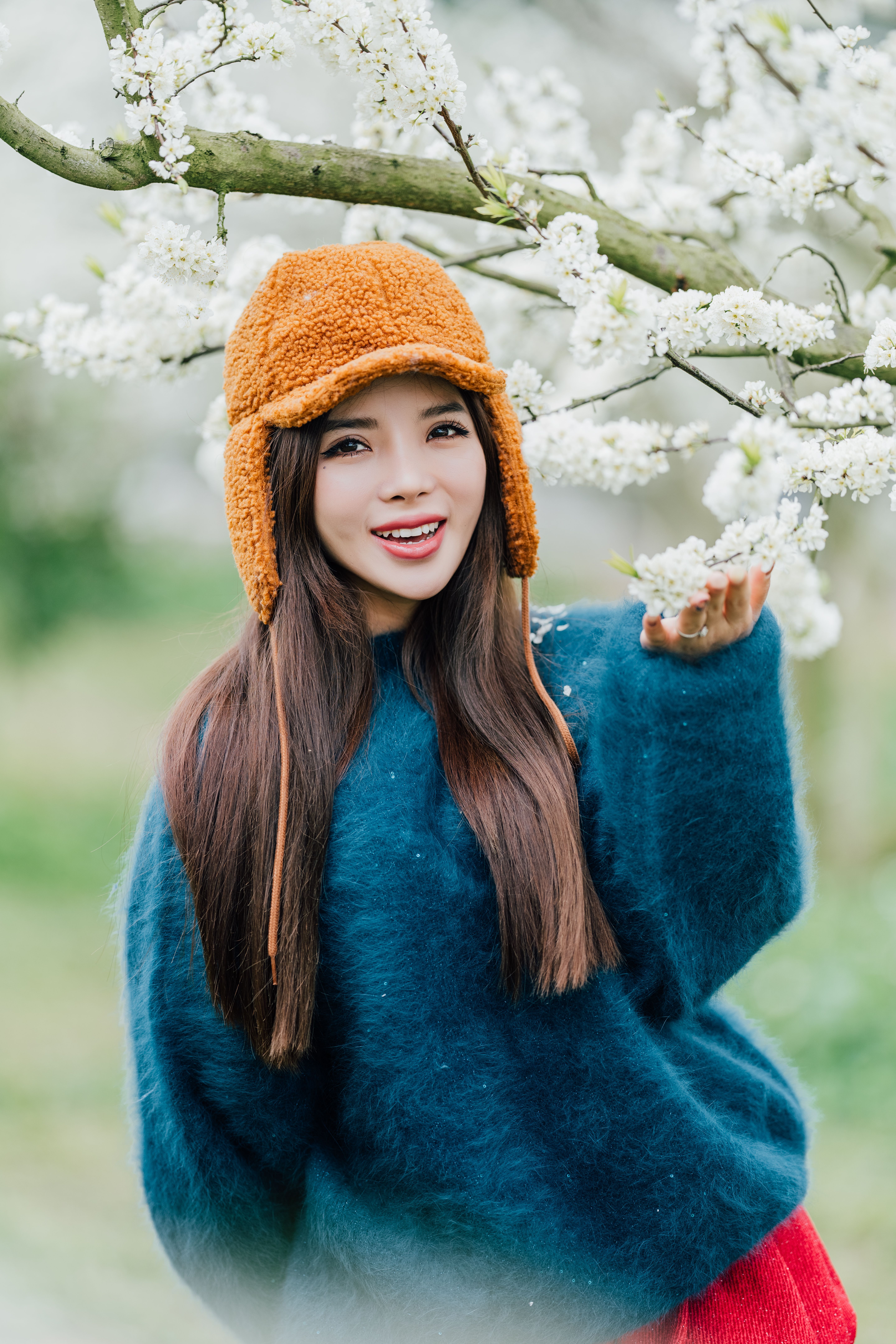 Cheerful woman in a warm hat and sweater appreciating spring blossoms in a garden.