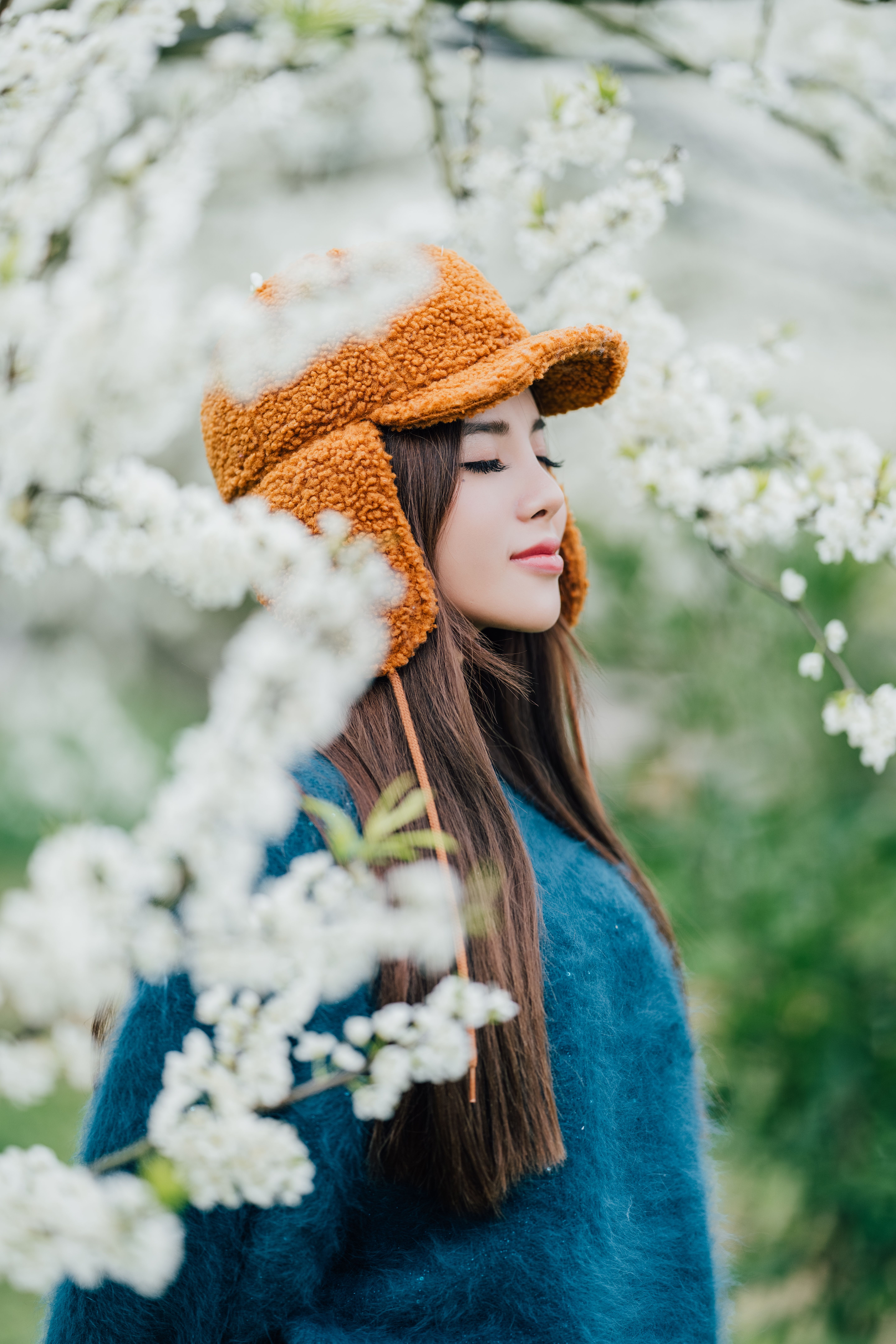 Young woman in a cozy orange hat amidst vibrant spring blossoms, eyes closed, enjoying nature.