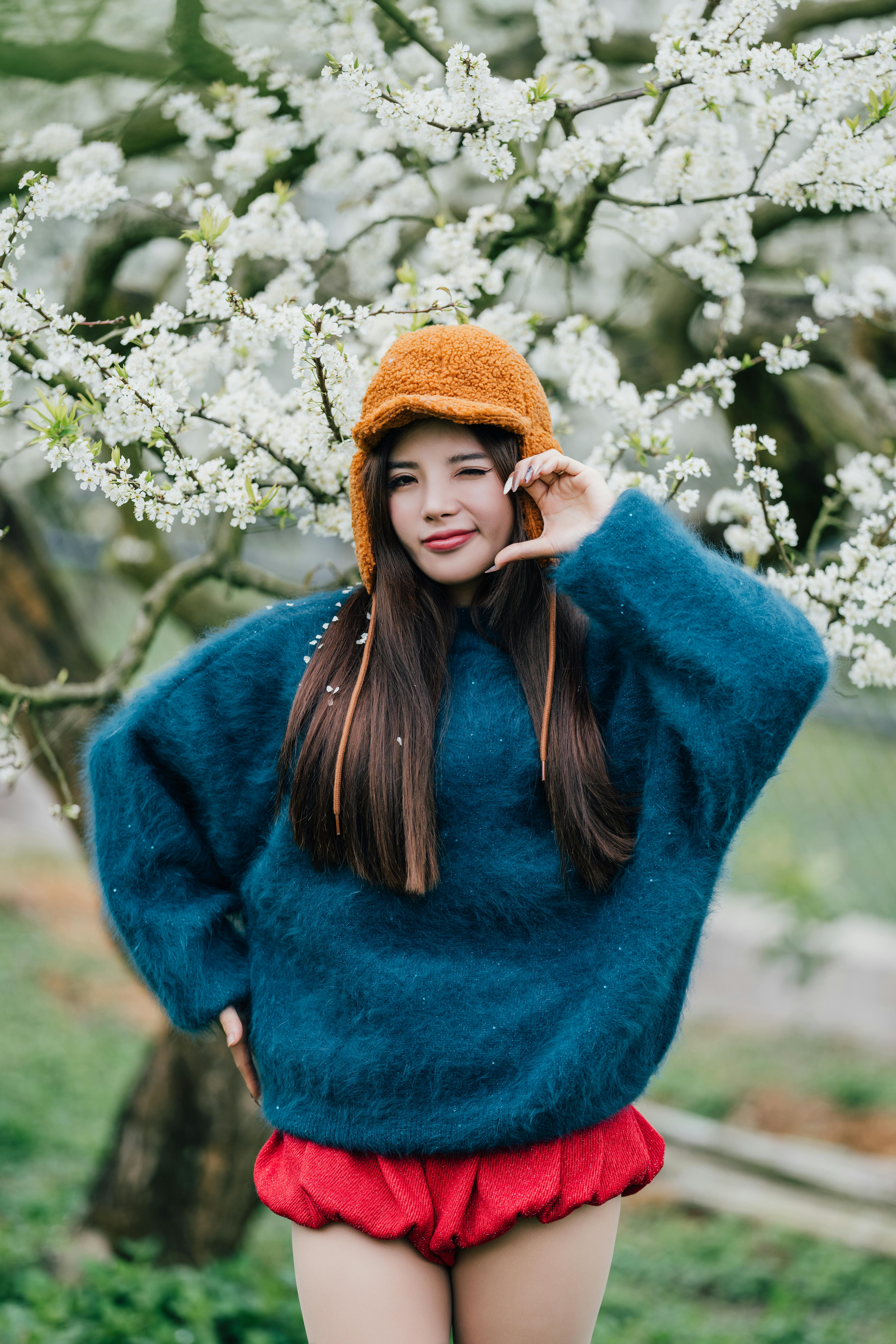 A smiling woman in a bright winter outfit poses in front of a blooming tree, capturing a joyful outdoor moment.