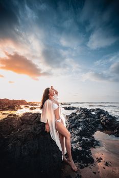 Model poses in white bikini and robe against beach rocks at sunset, capturing a serene and stylish moment.