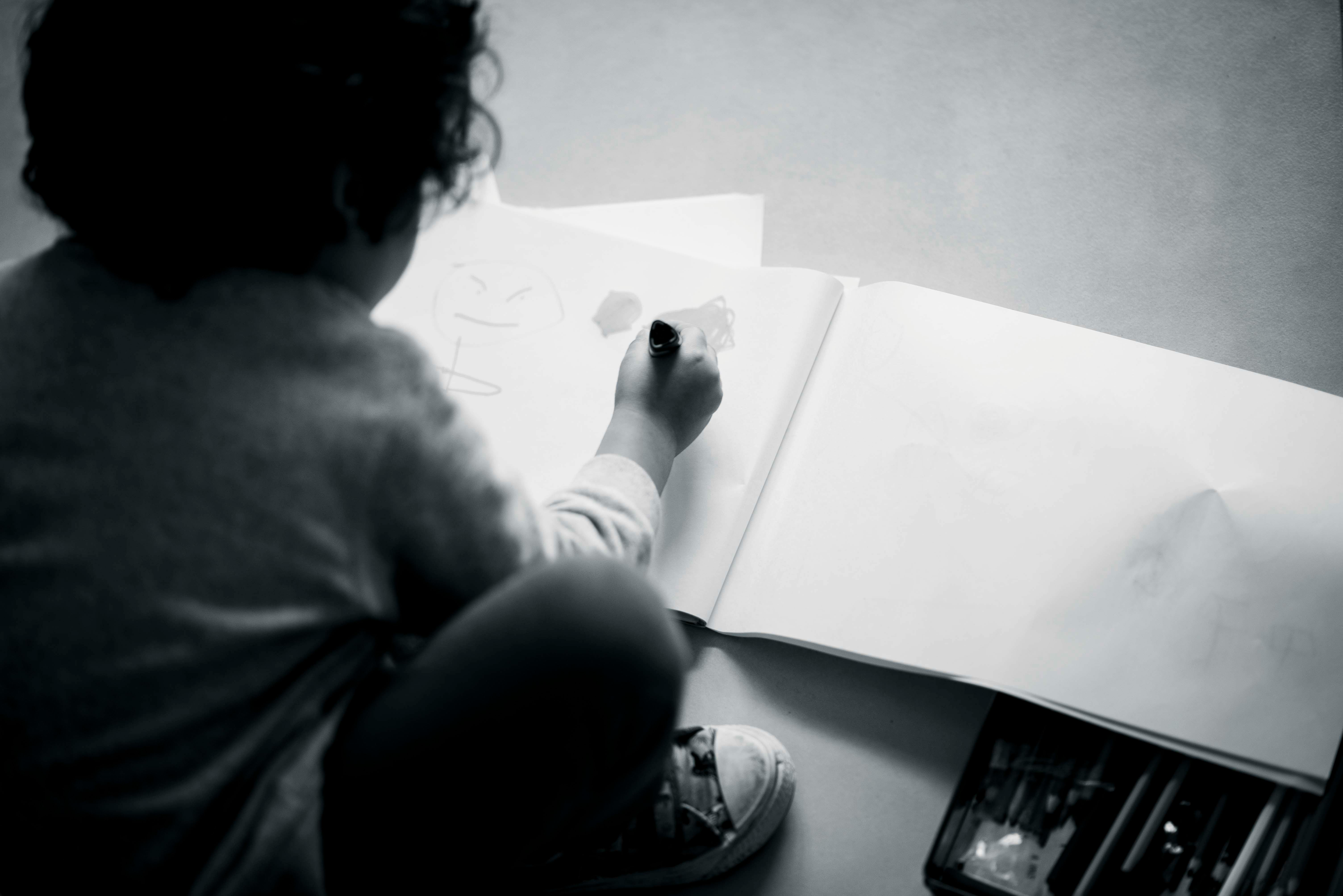 Gratis Un bambino che disegna su un grande blocco note con del materiale artistico a portata di mano, immortalato in bianco e nero. Foto a disposizione