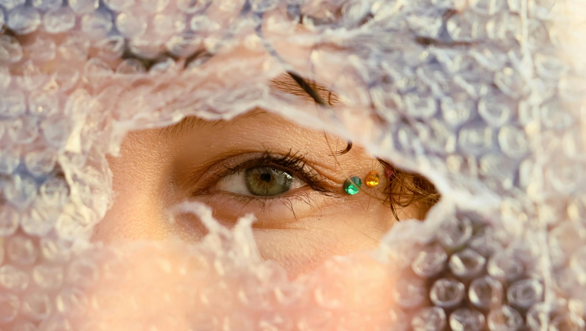 Close-up of a human eye peering through bubble wrap adorned with colored gems.