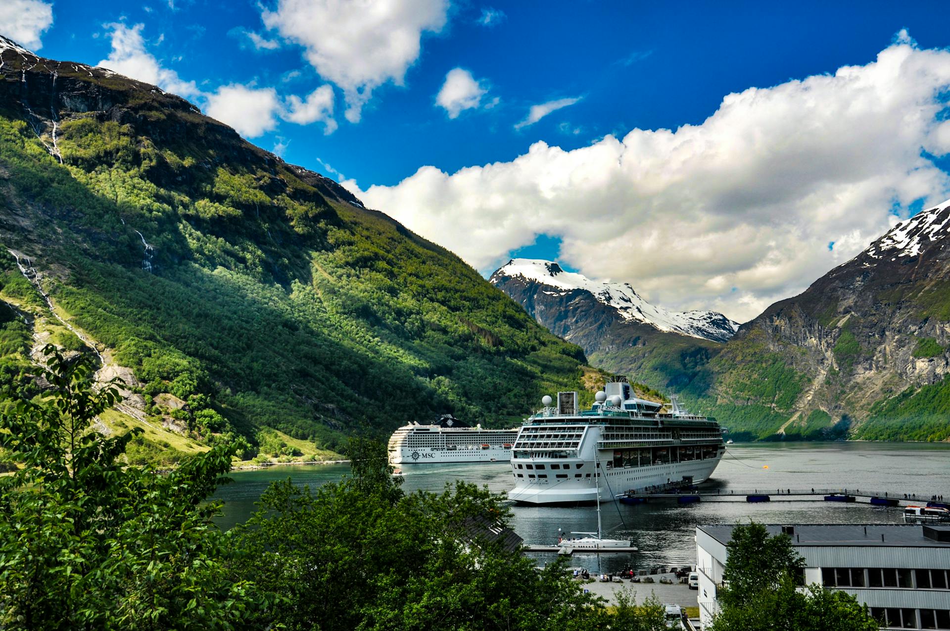 Cruise ship docked in picturesque Geiranger Fjord surrounded by lush mountains and clear blue sky.