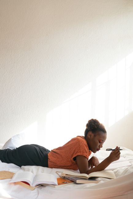 A young woman enjoys leisurely reading while lying on a bed in a sunlit room.