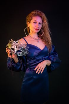 Stylish woman in blue satin dress holding a silver masquerade mask gracefully.