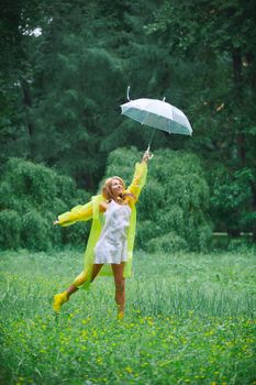 A woman joyfully leaps in a lush green field, holding an umbrella, wearing a yellow raincoat.