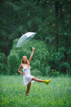 A woman in a white dress and yellow boots dances in lush greenery with an umbrella.