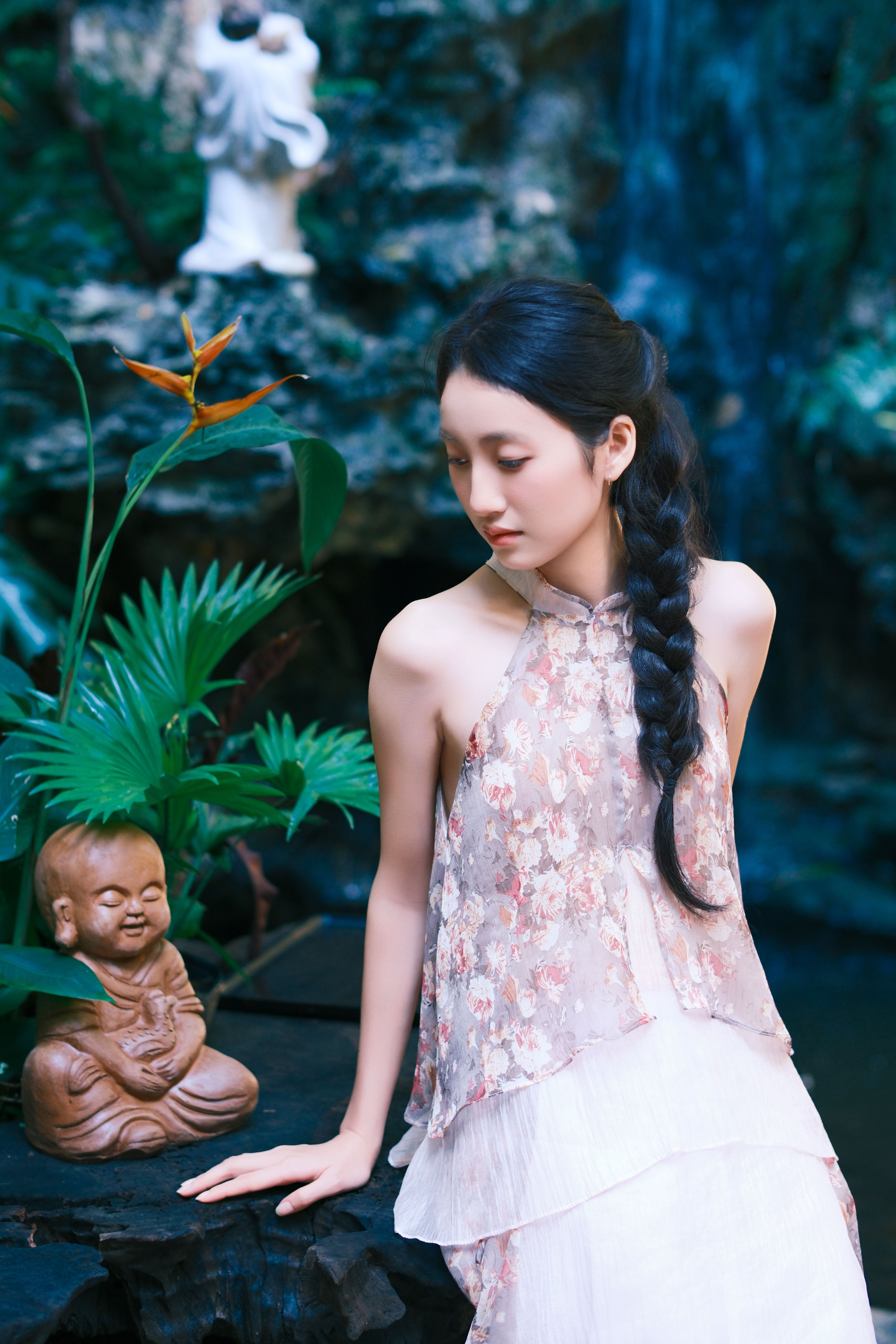 Woman in a floral dress meditates by a serene Buddha statue in a lush garden setting.