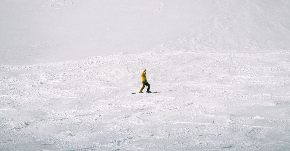 A solitary skier in a yellow jacket traverses a vast snowy mountain under a clear sky.