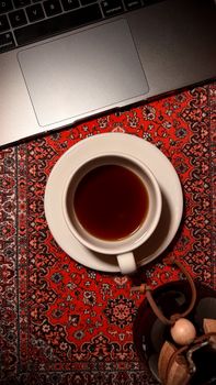 Cozy overhead shot of a tea cup on a decorative oriental tablecloth beside a laptop.