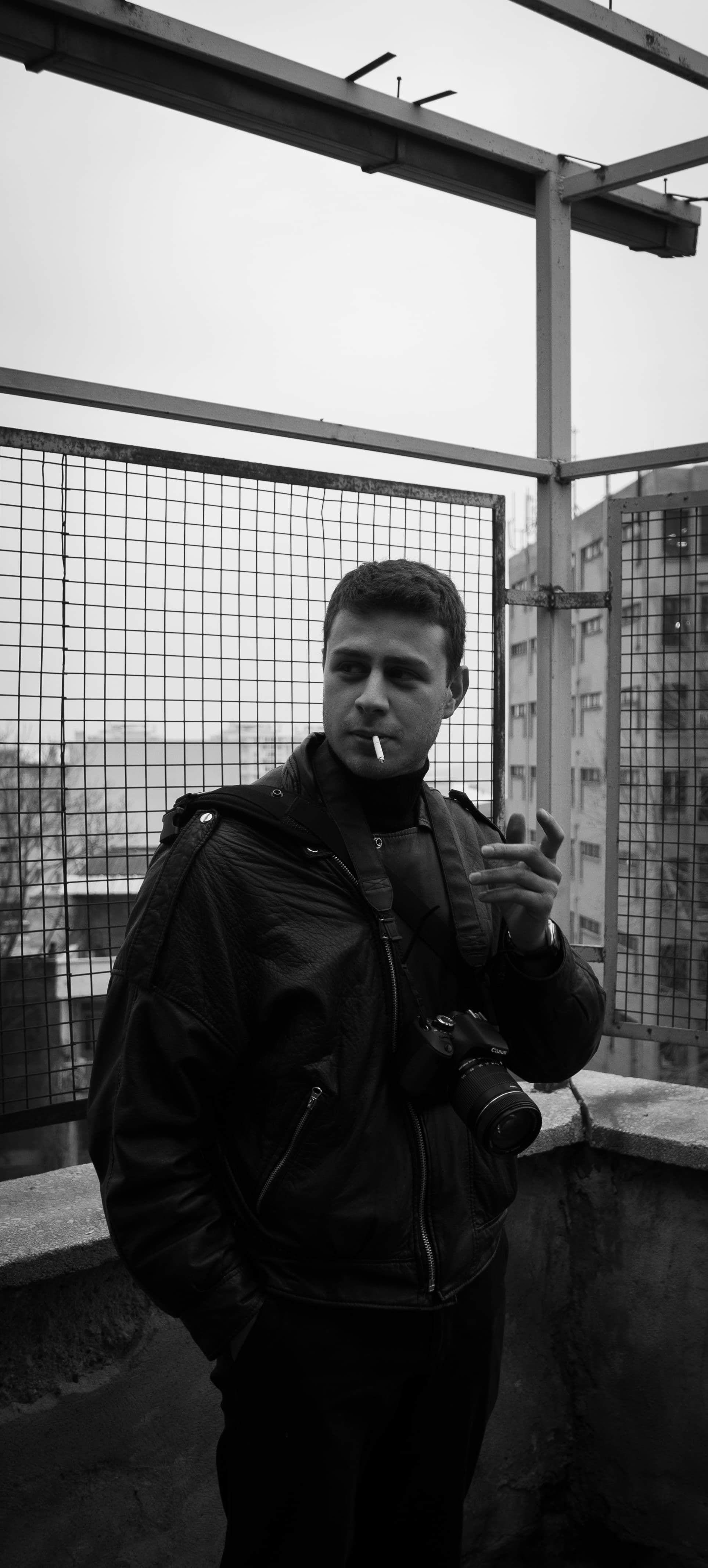 A photographer with a camera stands smoking, in a black and white urban setting.