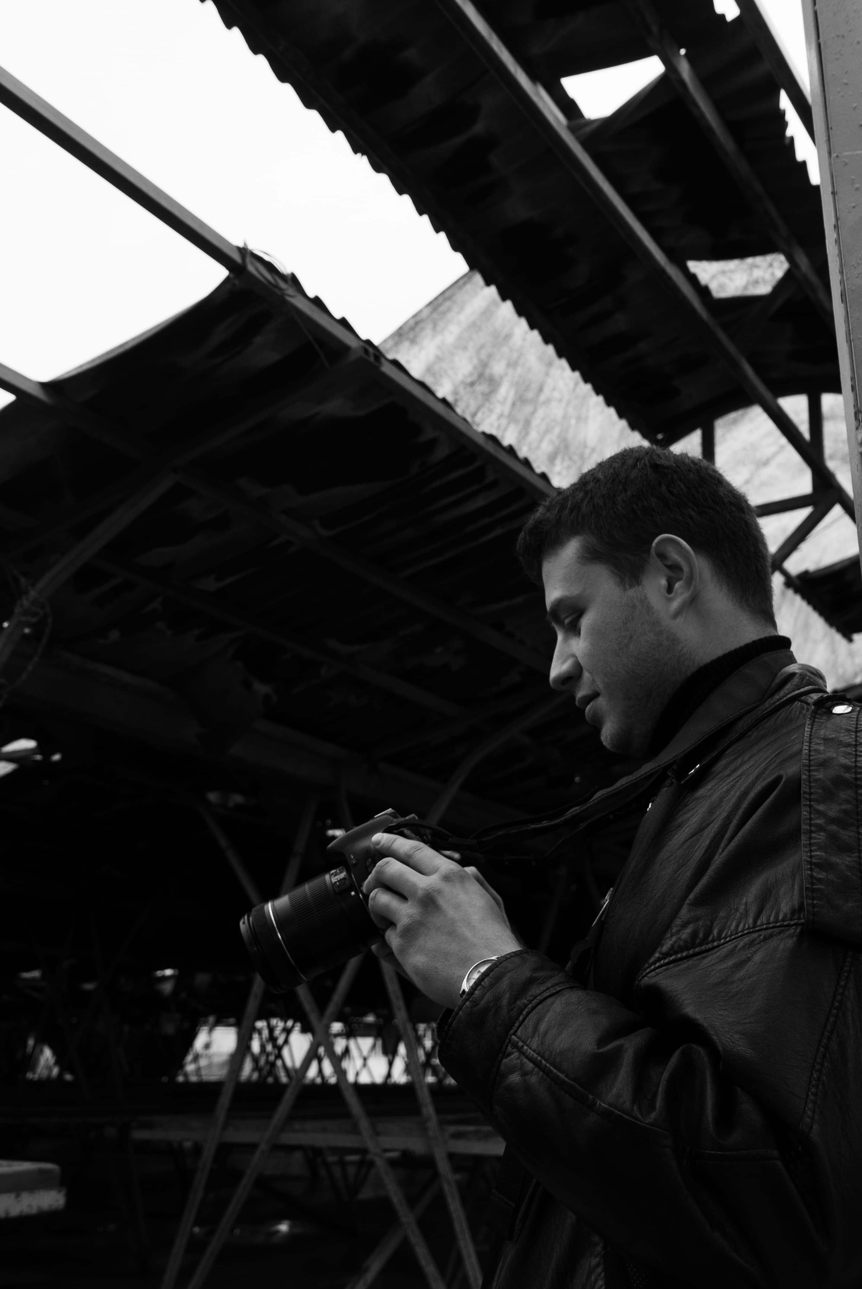 A man in leather jacket examines his camera in an industrial setting.