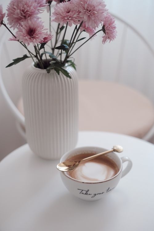 Free A cup of coffee with a gold spoon next to pink flowers in a vase on a white table. Stock Photo