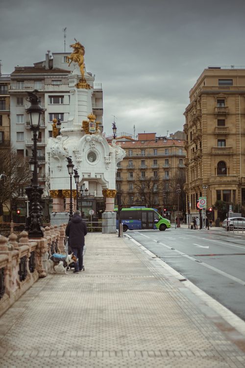 Free Pedestrian walking a dog on a city street with ornate historic architecture and urban scenery. Stock Photo