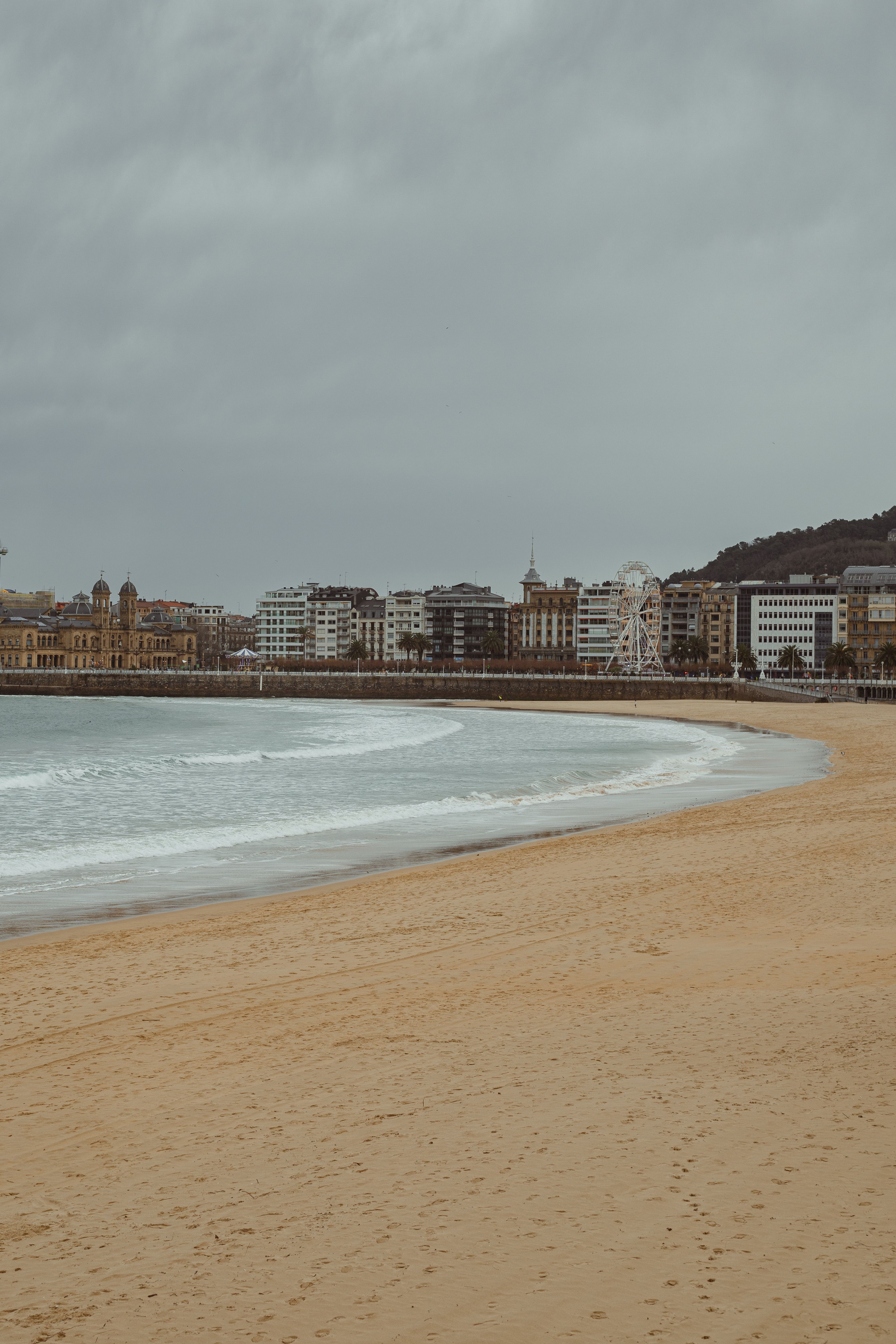 San Sebastián's beach with an overcast sky, cityscape and calm waves create a serene coastal scene.