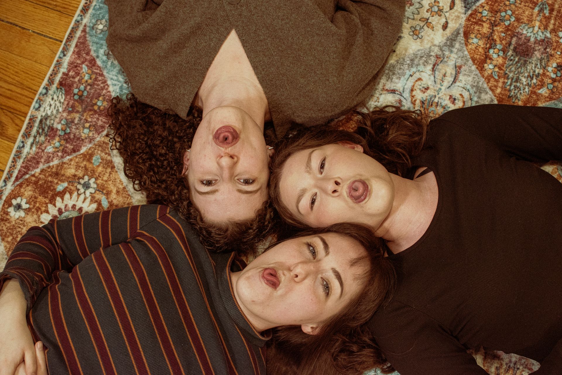 Three adult friends lying on a patterned carpet, making funny faces together.
