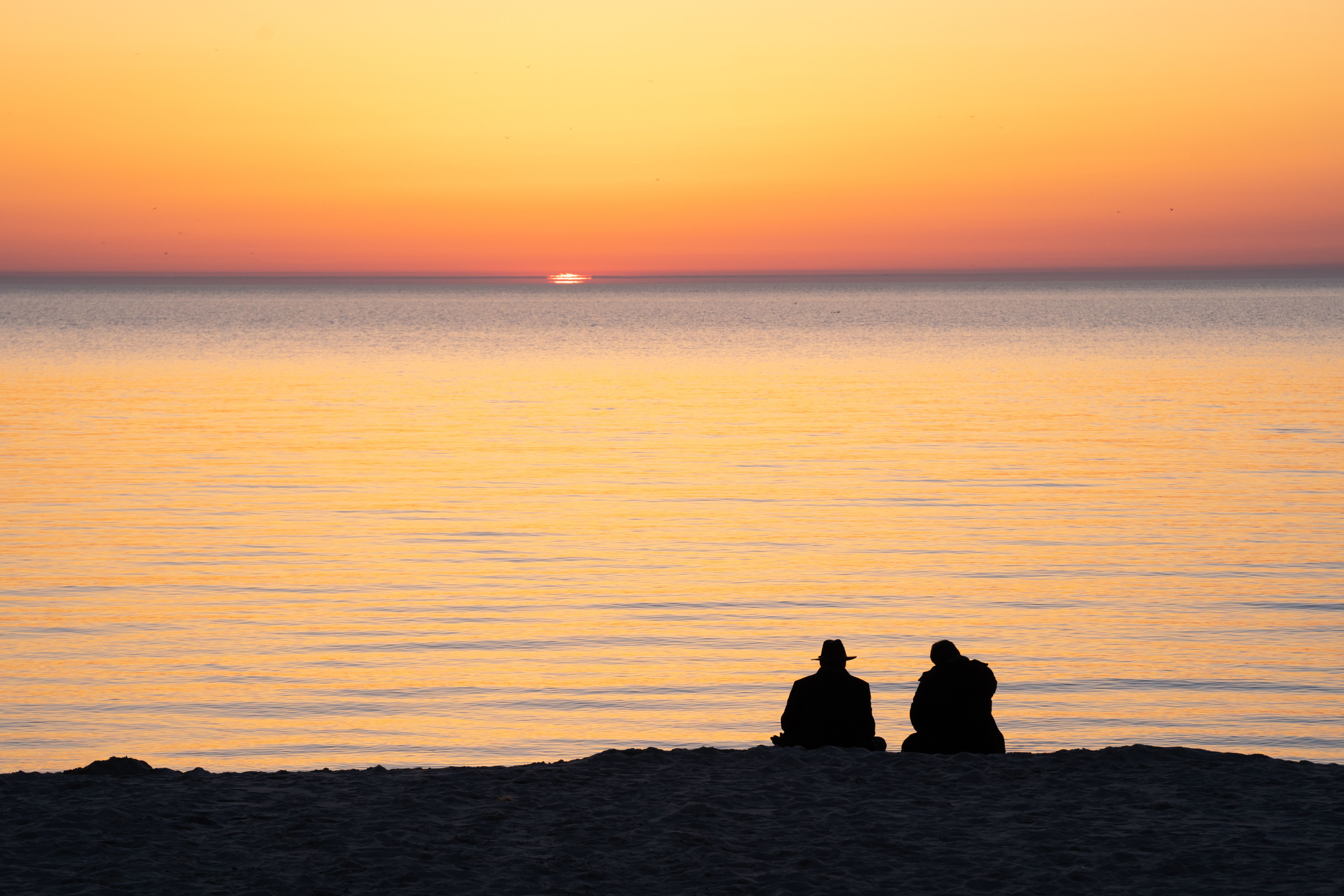 Silhouette of two people sitting on a beach during a vibrant sunset over calm waters.