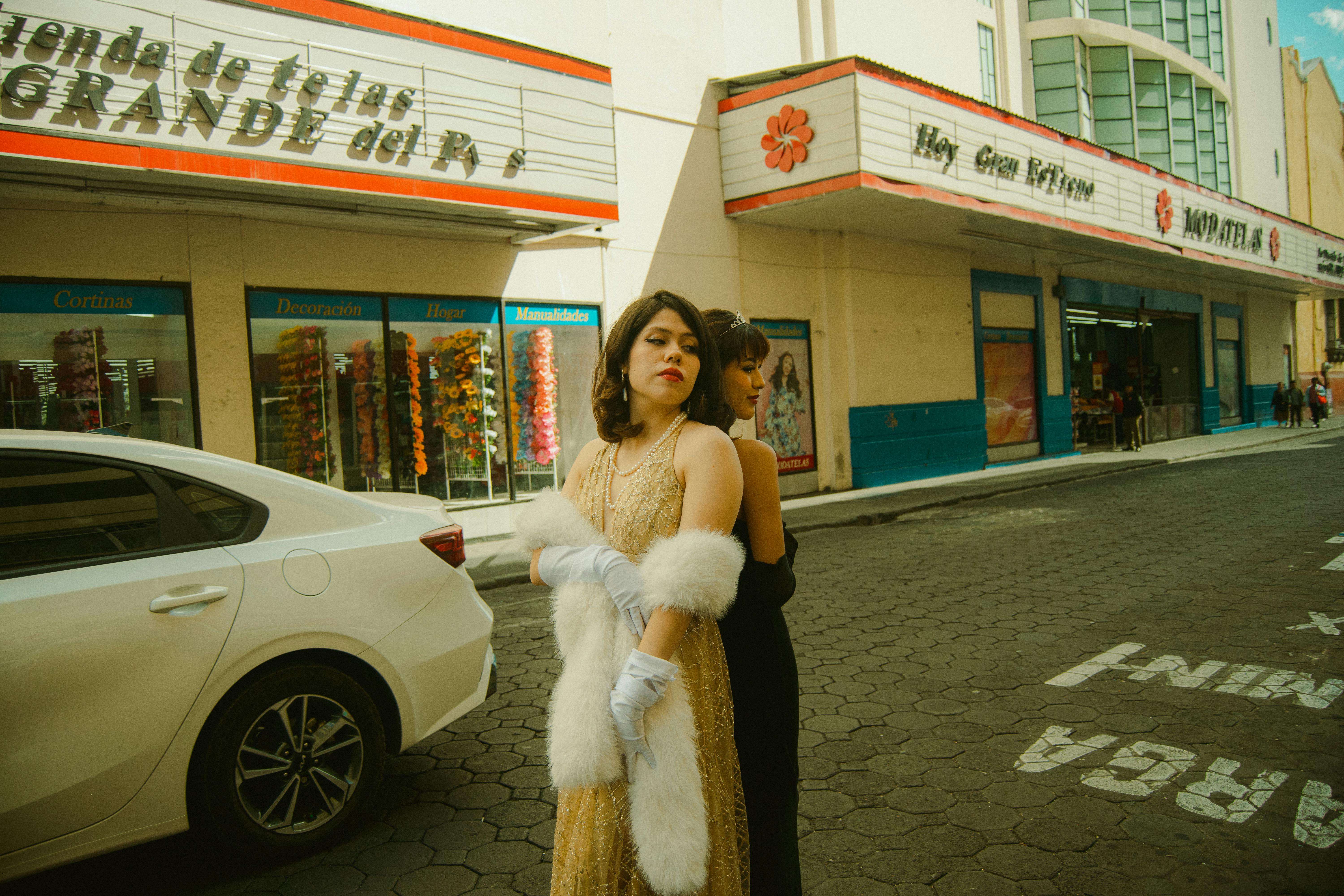 Women in elegant vintage attire pose in front of an urban street with retro signage.