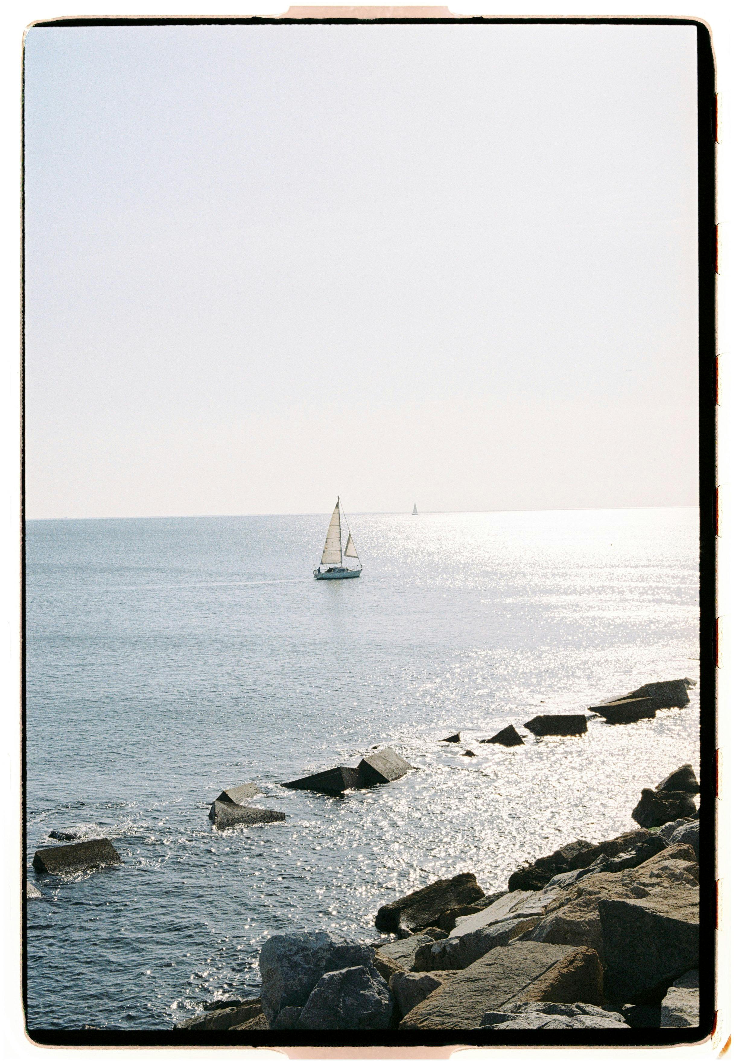 A tranquil scene of a sailboat gliding on a calm sea near a rocky shoreline.