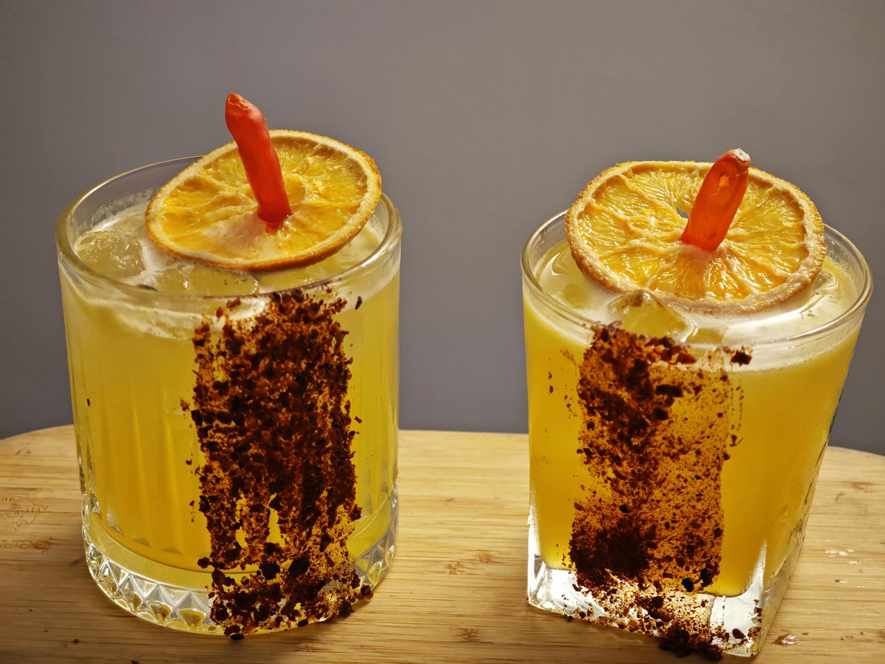 Two vibrant citrus cocktails garnished with dried orange slices and chili, perfect for spicy drink lovers.