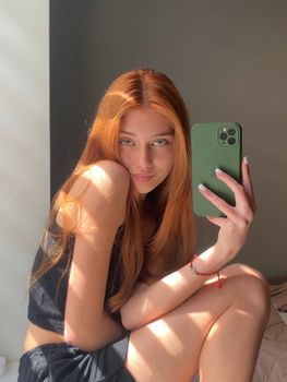 Portrait of a young woman with long red hair taking a selfie in bright sunlight.