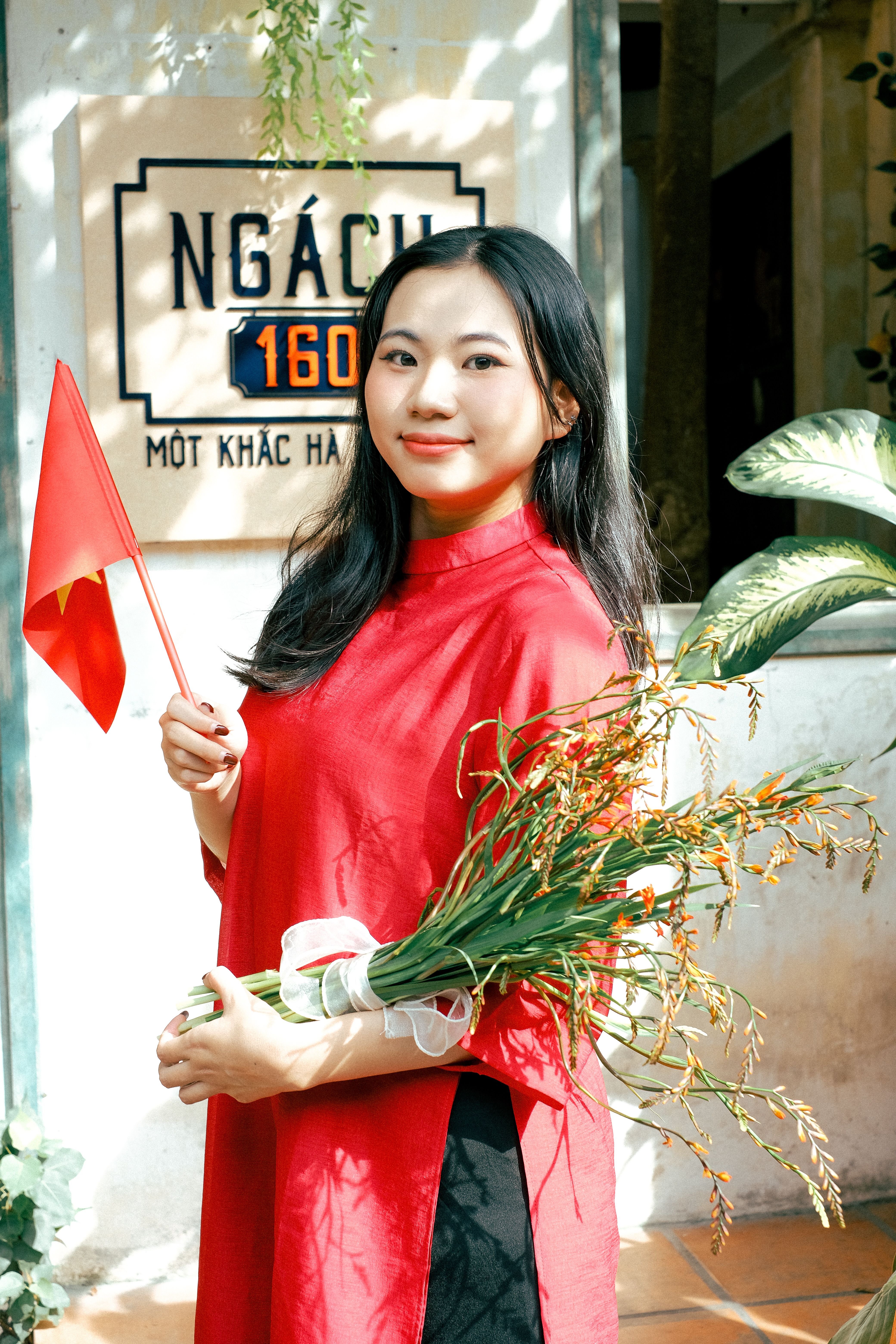Smiling woman in red dress holds bouquet and flag, outdoor setting with sign.