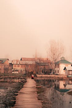 A peaceful village scene with a wooden path over water, leading to rustic houses surrounded by bare trees.