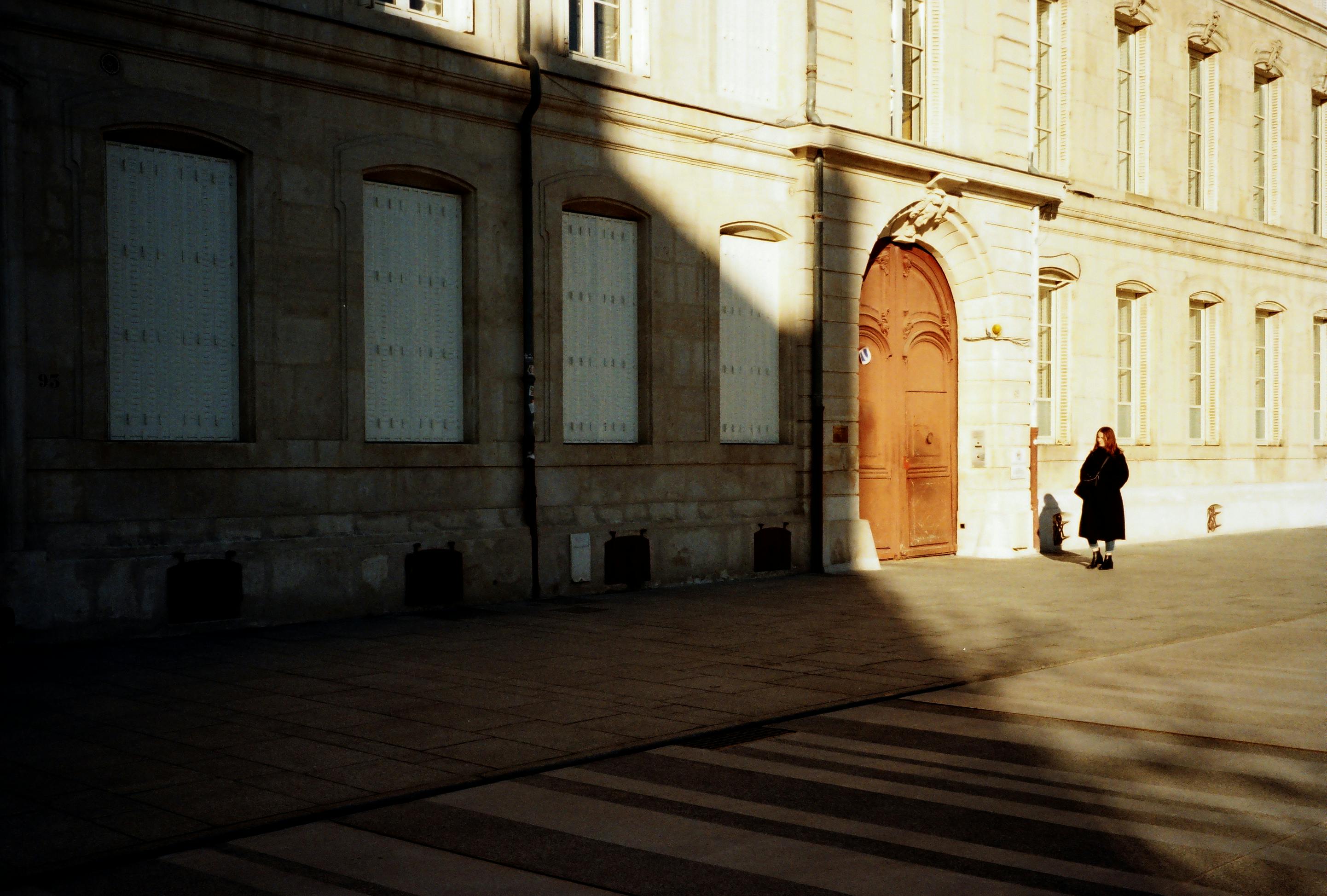 A person walks past a historic building with intriguing shadow patterns on a sunny day.