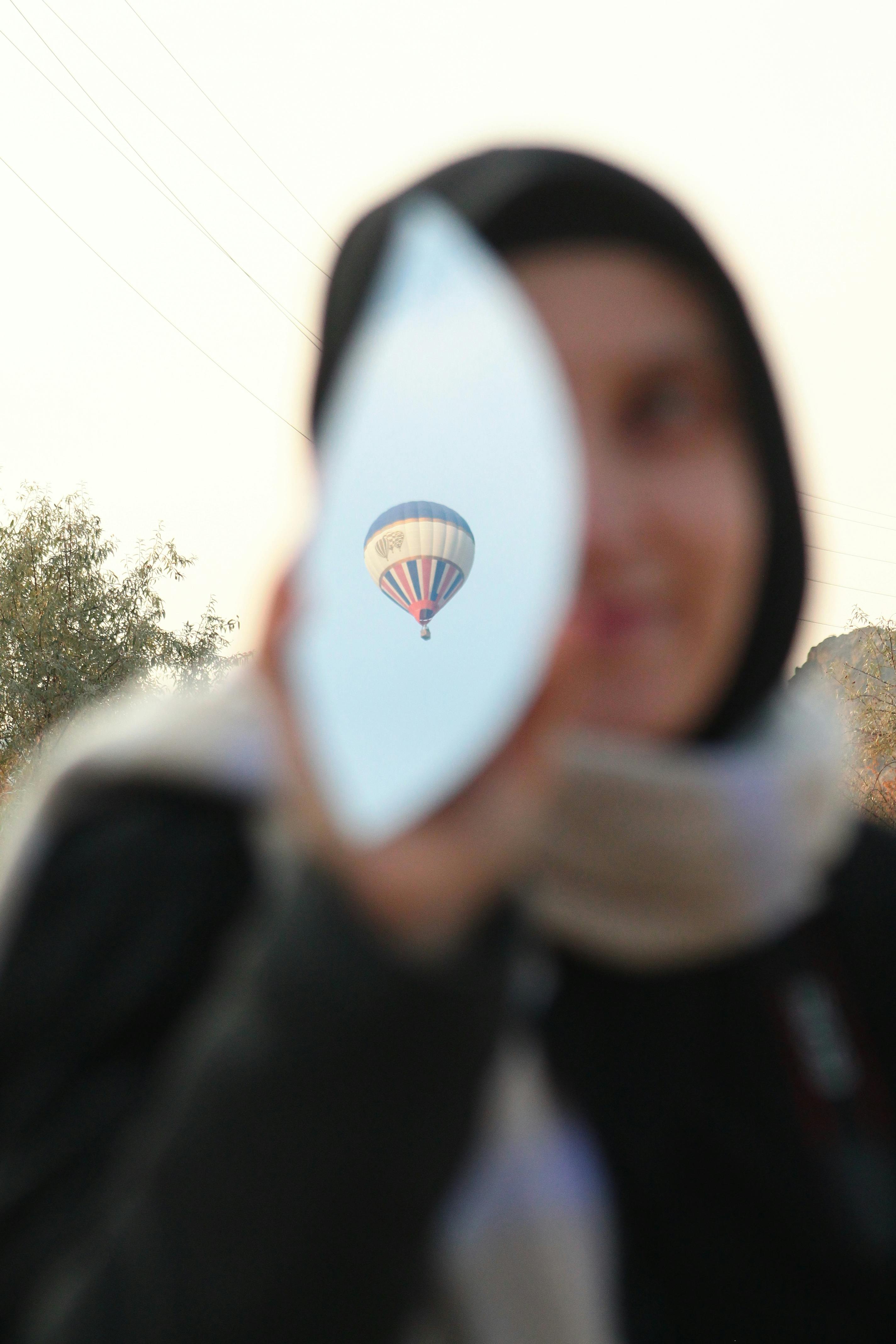 Hot air balloon reflected in a mirror held by a woman in a blurred background.