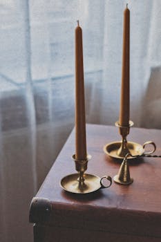 Warm scene with brass candle holders on wooden furniture by a window.