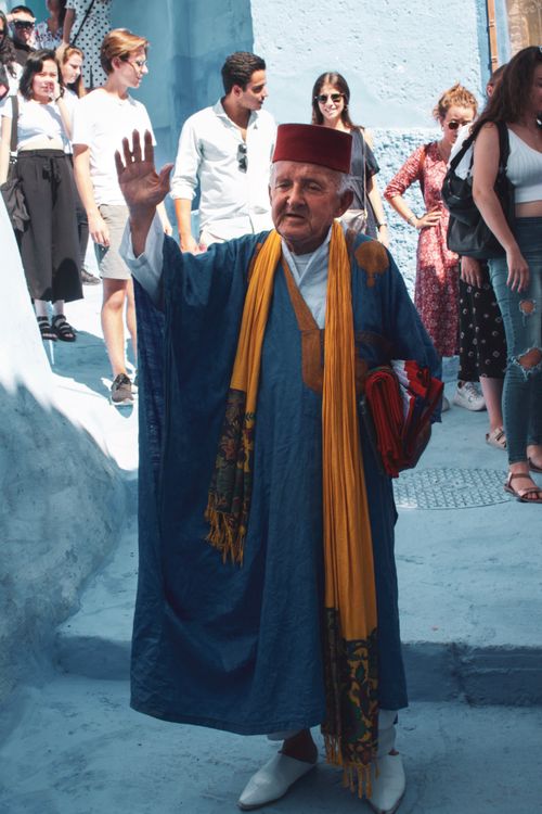Free An elder in traditional Moroccan clothing waves in a bustling Chefchaouen street. Stock Photo