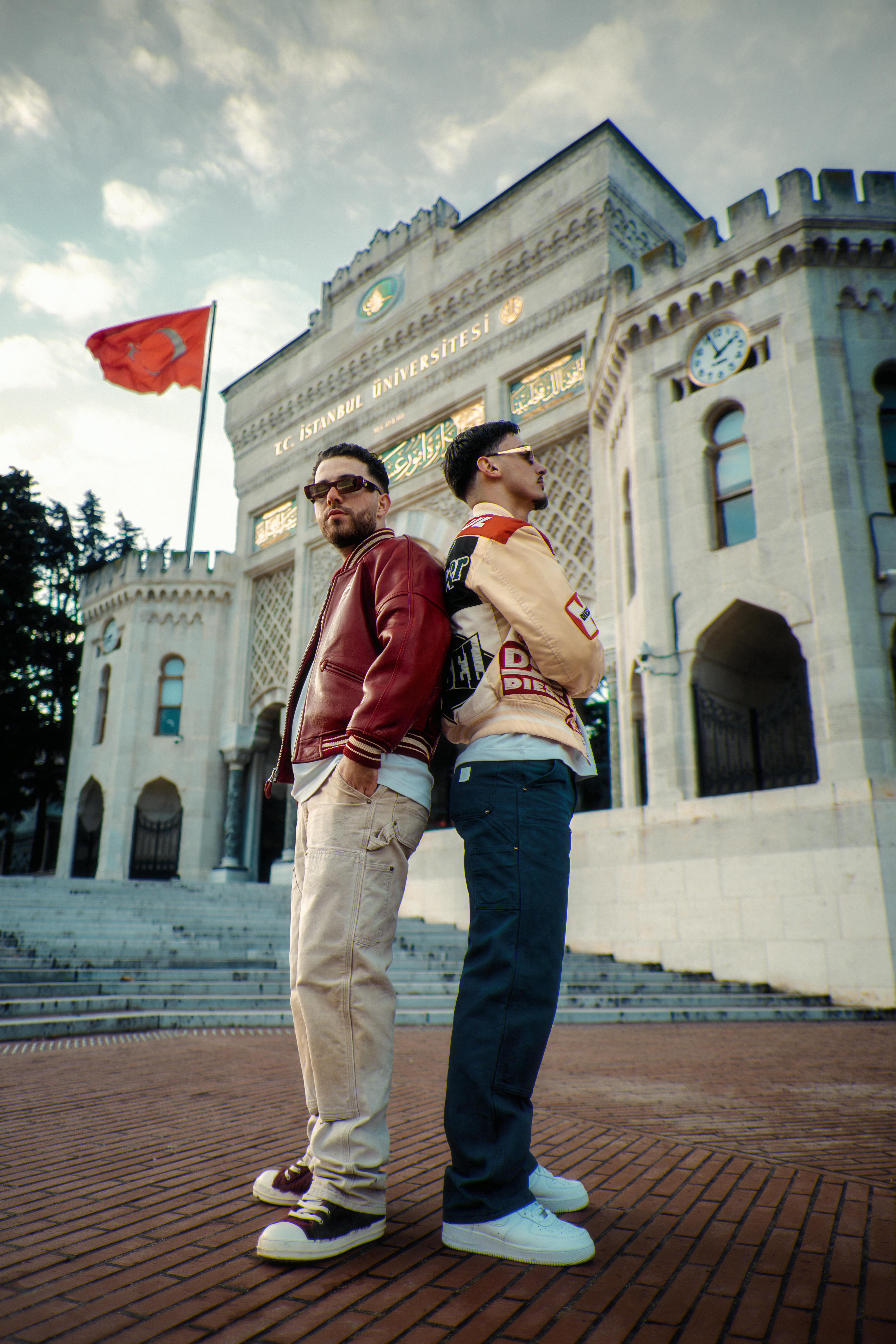 Two fashionable men in casual attire pose at a historic university entrance.