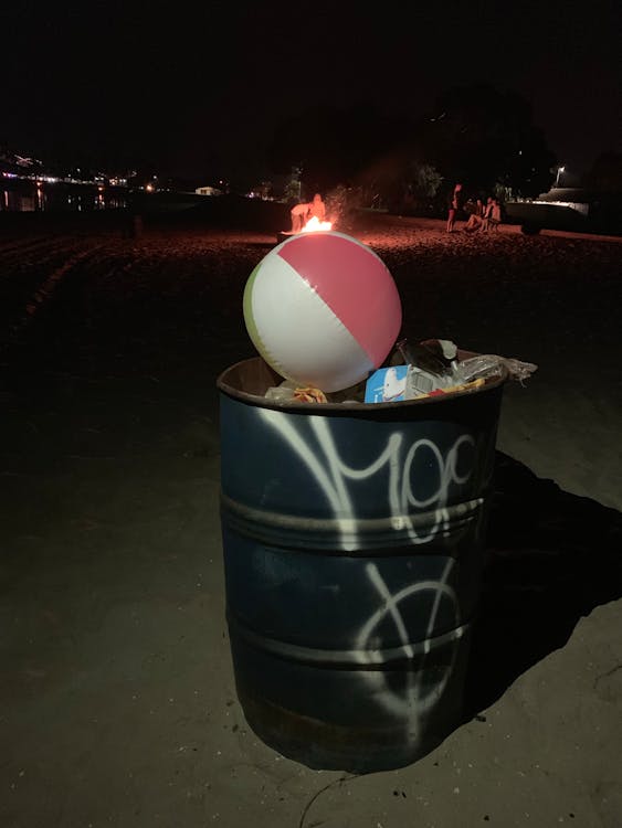 Garbage container filled with rubbish and beach ball placed on coast against burning campfire
