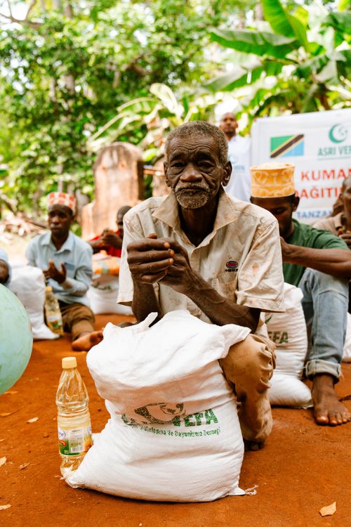 Free An elderly man sits outdoors with donated food supplies, surrounded by community. Stock Photo