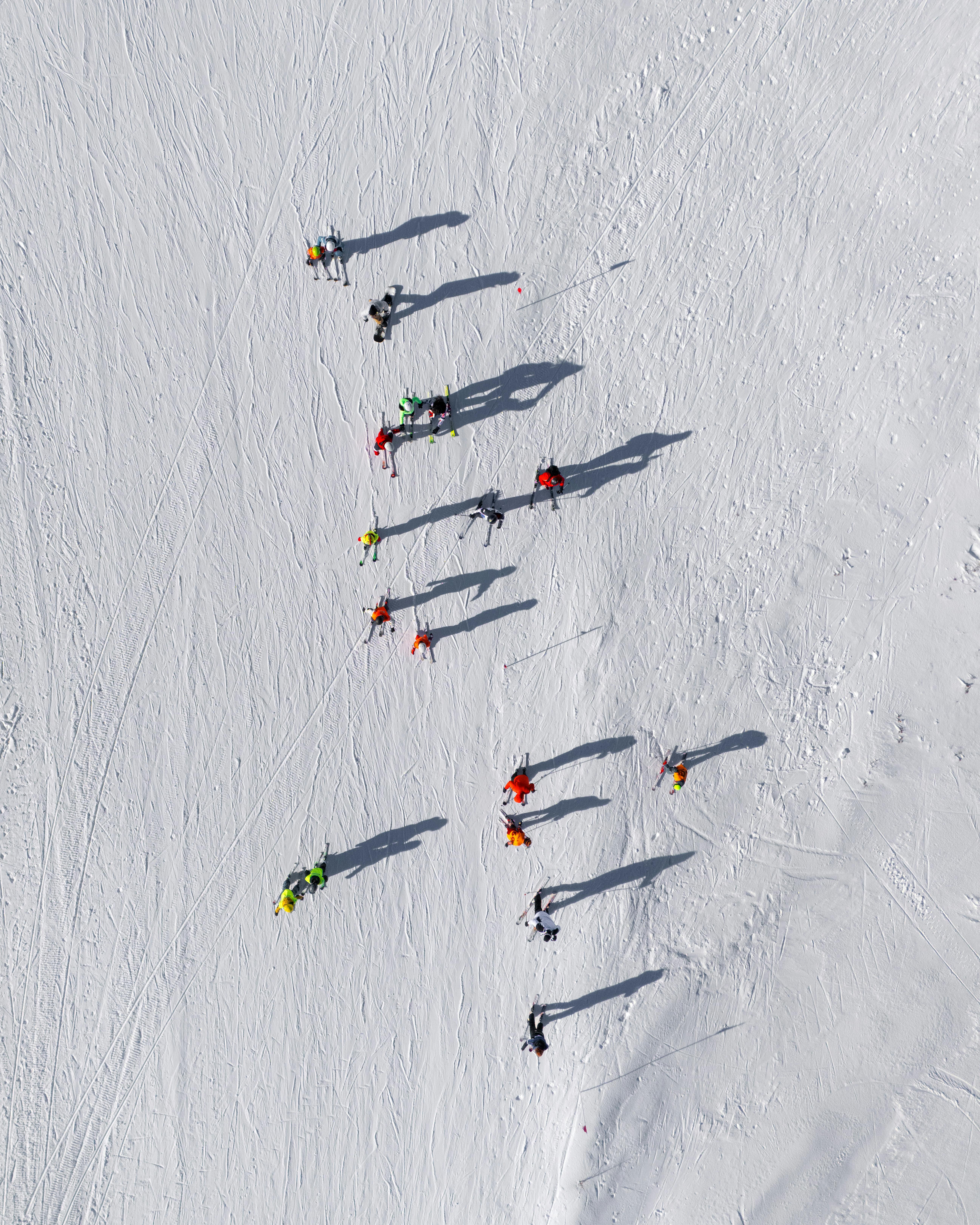 Aerial shot of skiers on a snowy slope casting long shadows, with bright ski apparel.