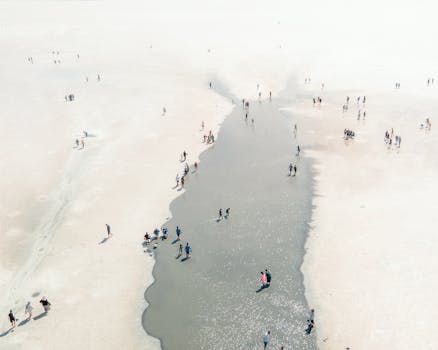 People walking along a narrow water stream in a vast sandy landscape, viewed from above.
