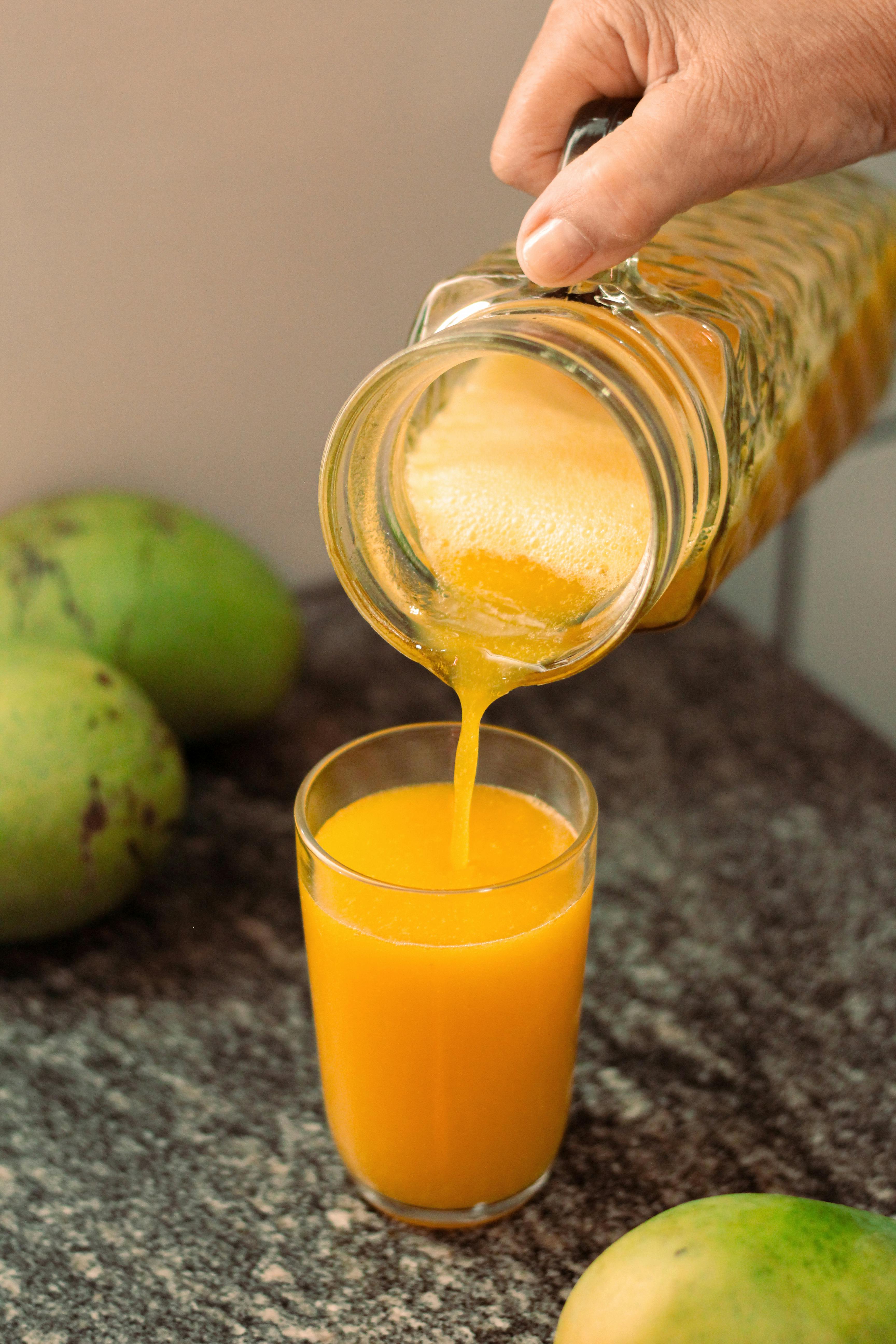 A refreshing glass of mango juice being poured from a jug, with fresh mangoes on the side.