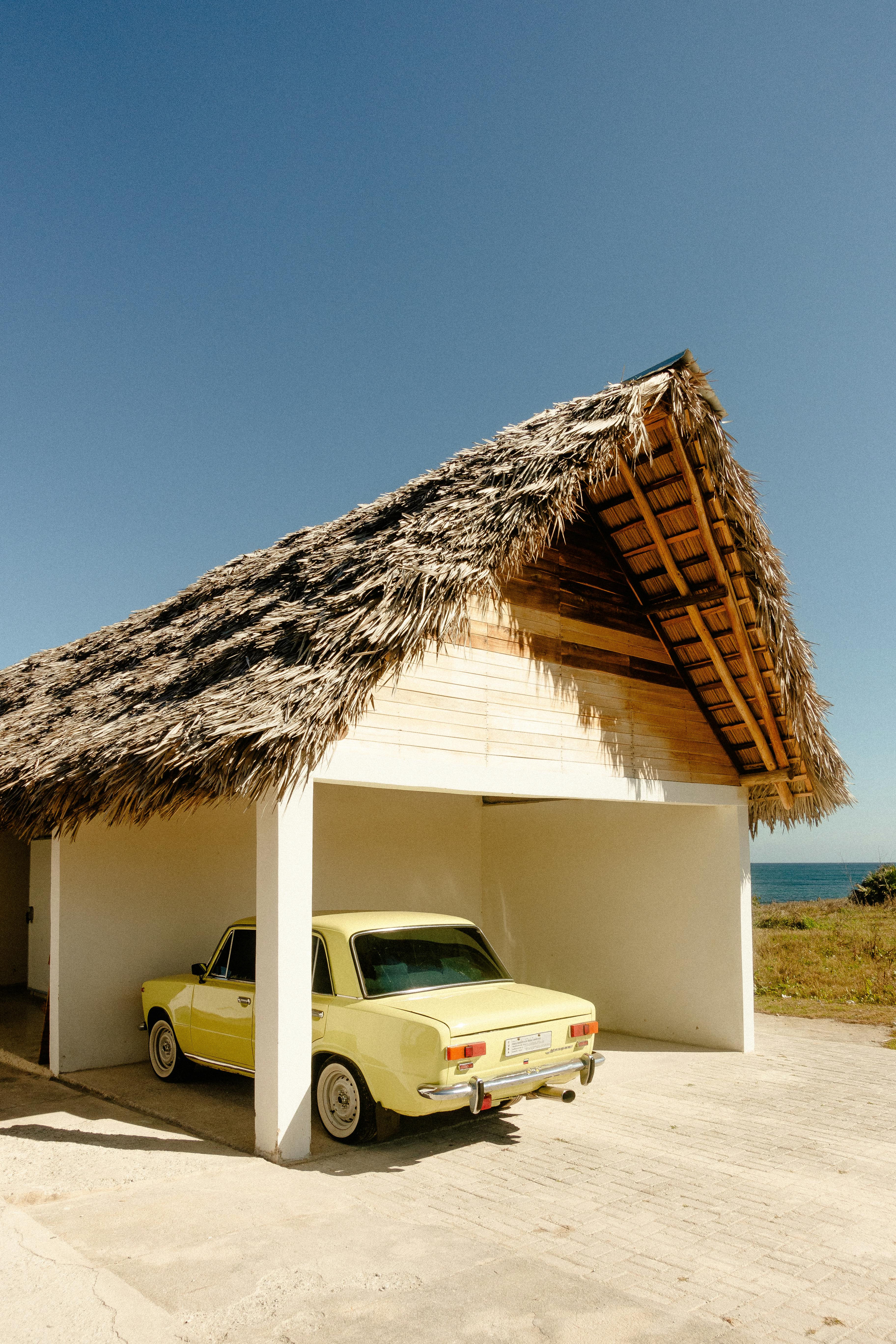 A classic yellow car parked under a thatched roof hut by the seaside.