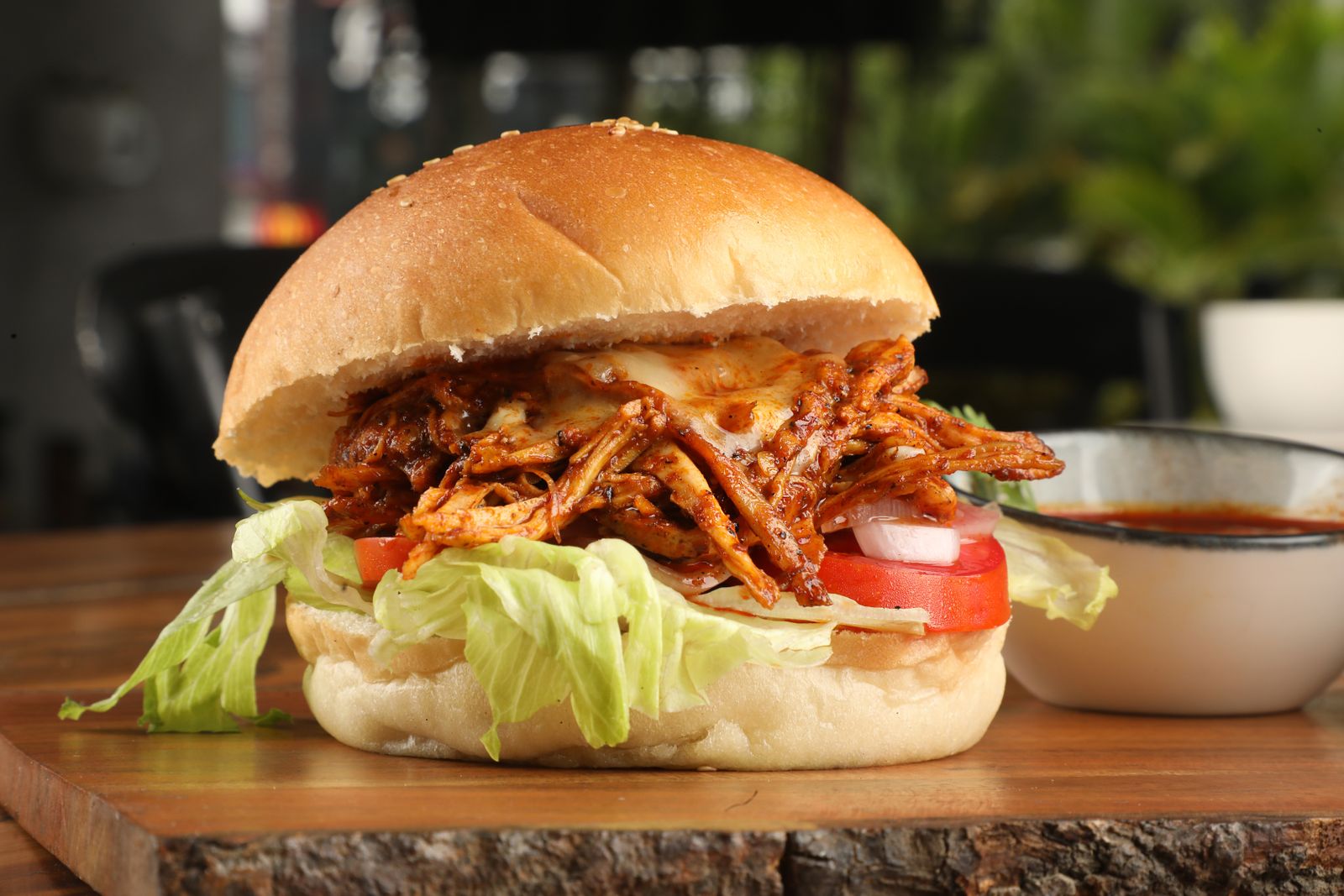 Pulled chicken burger with lettuce and tomato on a wooden board, with a bowl of sauce alongside.