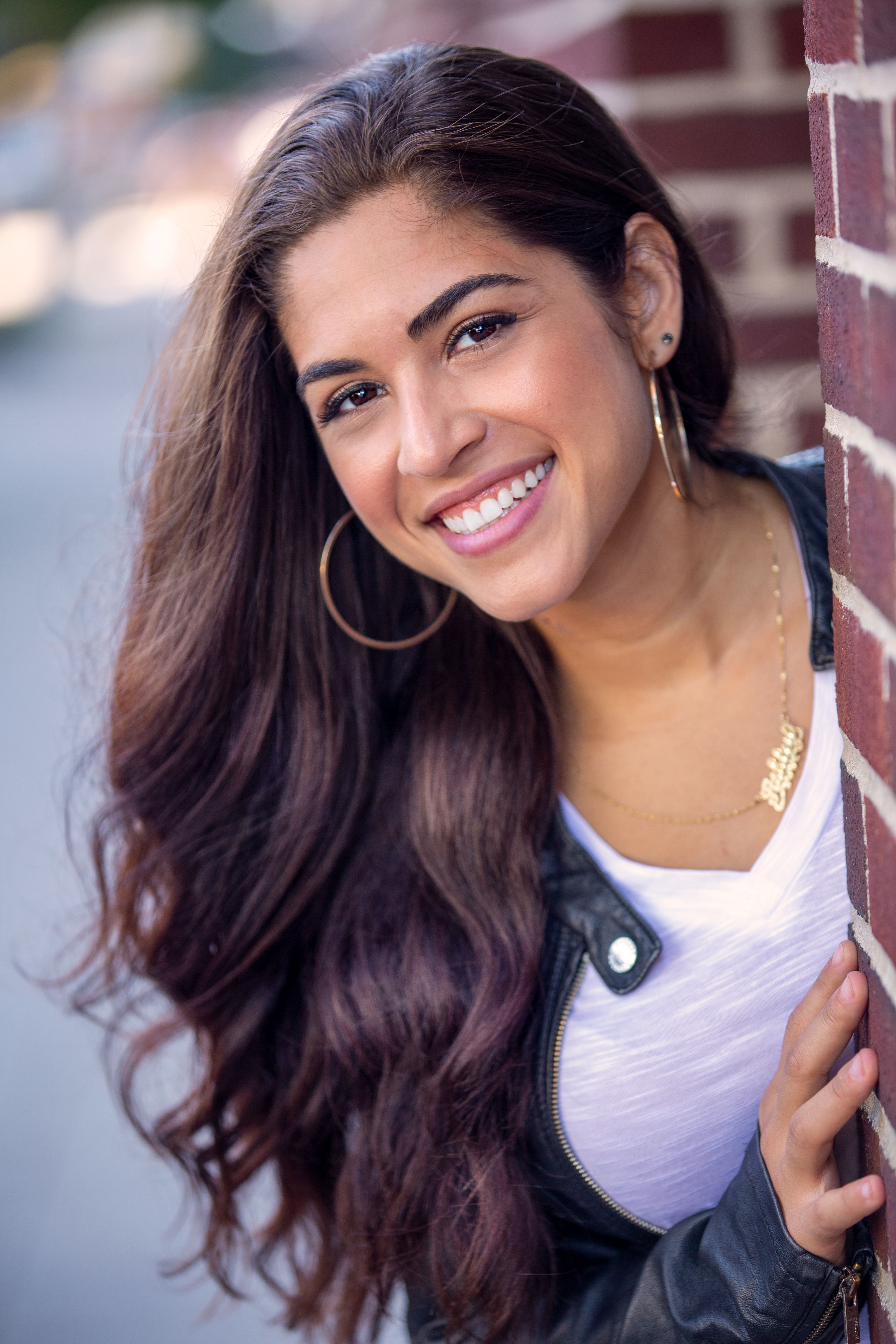 Portrait of a smiling woman with long hair leaning on a brick wall outdoors.