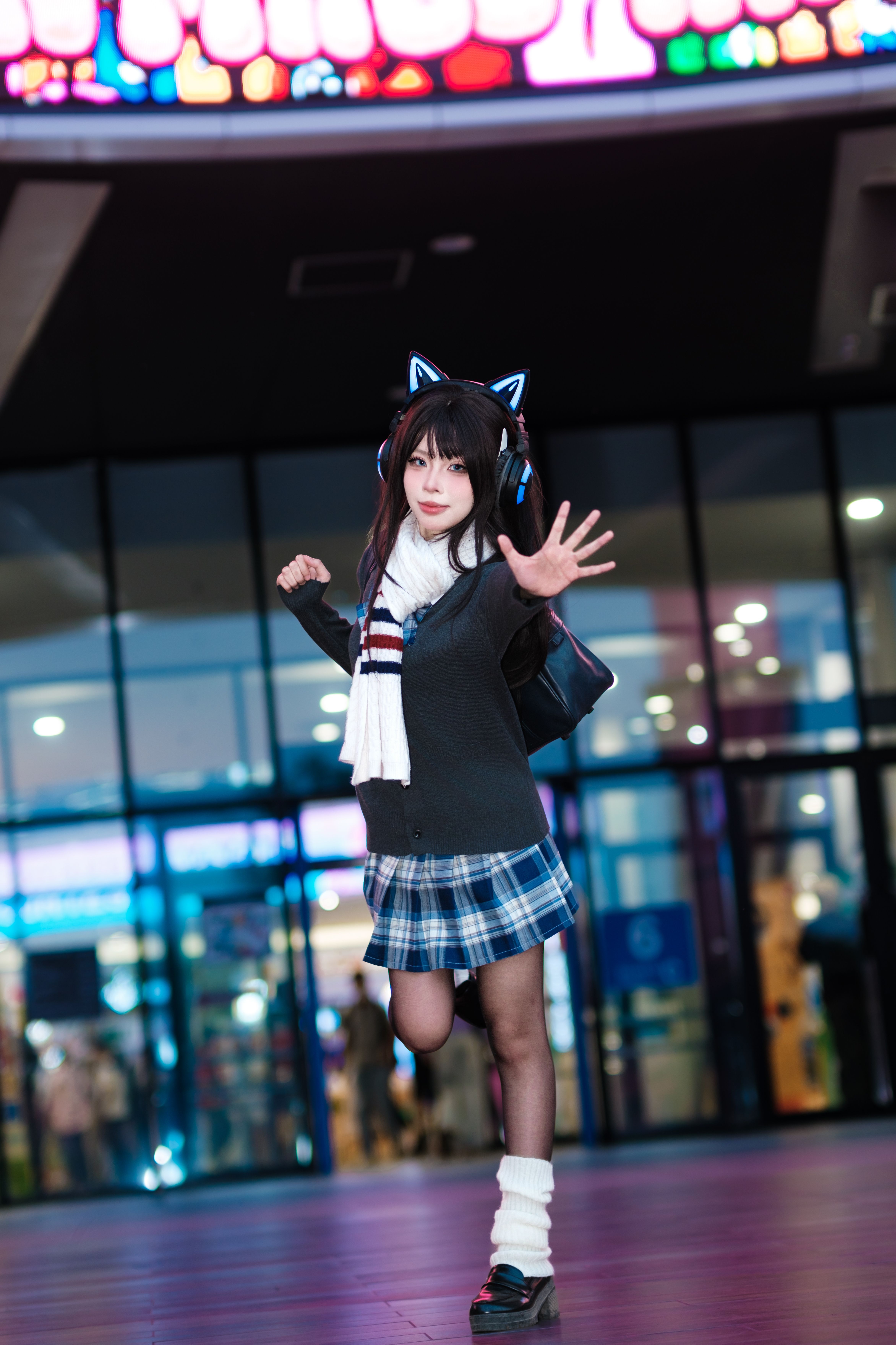 Cosplayer in cat-ear headphones and plaid skirt posing energetically in bustling city scene.