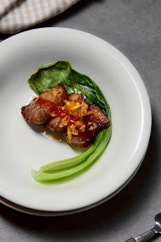 Top view of a gourmet pork belly dish with garnishing vegetables on a white plate.