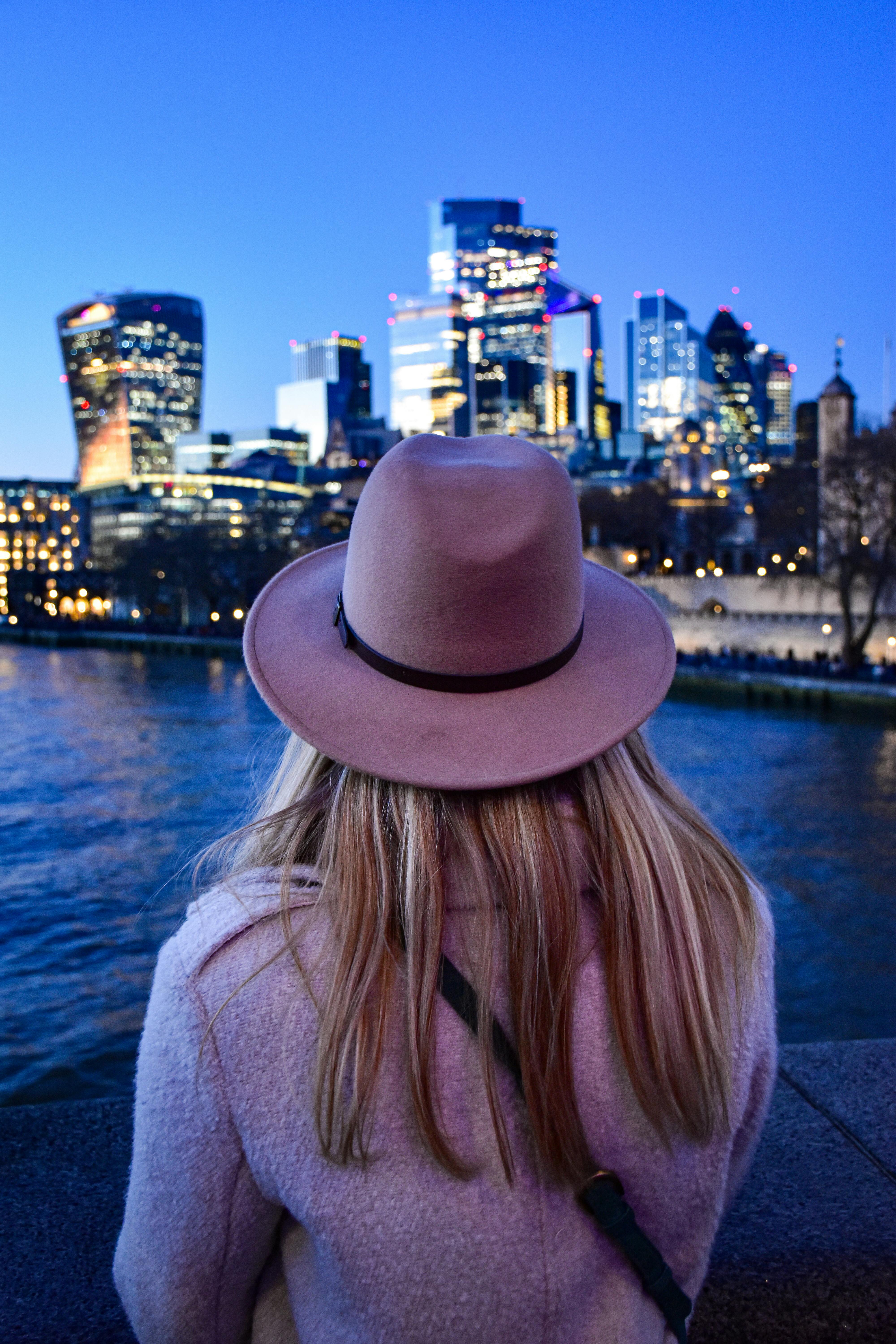 A woman in a hat observes the stunning London skyline at twilight with city lights glowing softly.