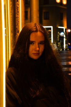 A young woman gazes pensively outdoors at night, with illuminated buildings providing a moody backdrop.