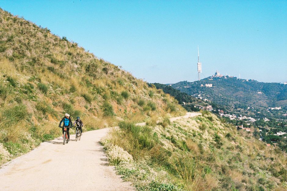 Cyclists ride along a mountain trail with distant views of a prominent TV tower.