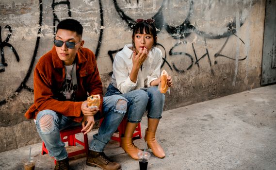 Young adults in casual attire eating street food by a graffiti wall, showcasing urban lifestyle.