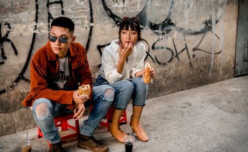 Free Young adults in casual attire eating street food by a graffiti wall, showcasing urban lifestyle. Stock Photo