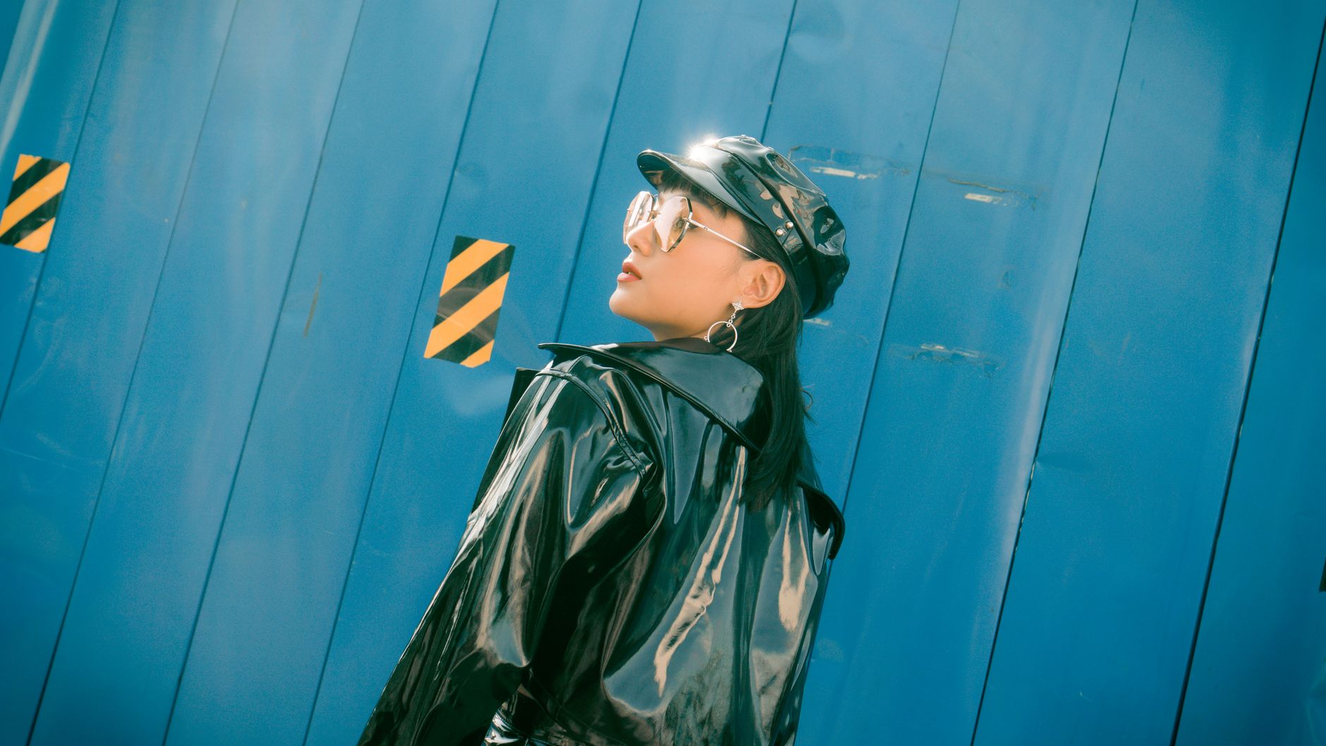 A stylish woman in a glossy black jacket posing outdoors against a vibrant blue industrial background.